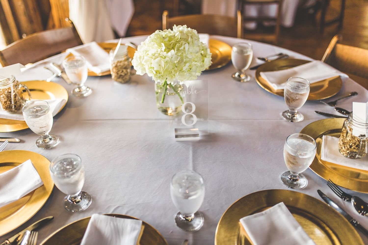 Elegant table setting with white linens, gold chargers, and a centerpiece of white hydrangeas.