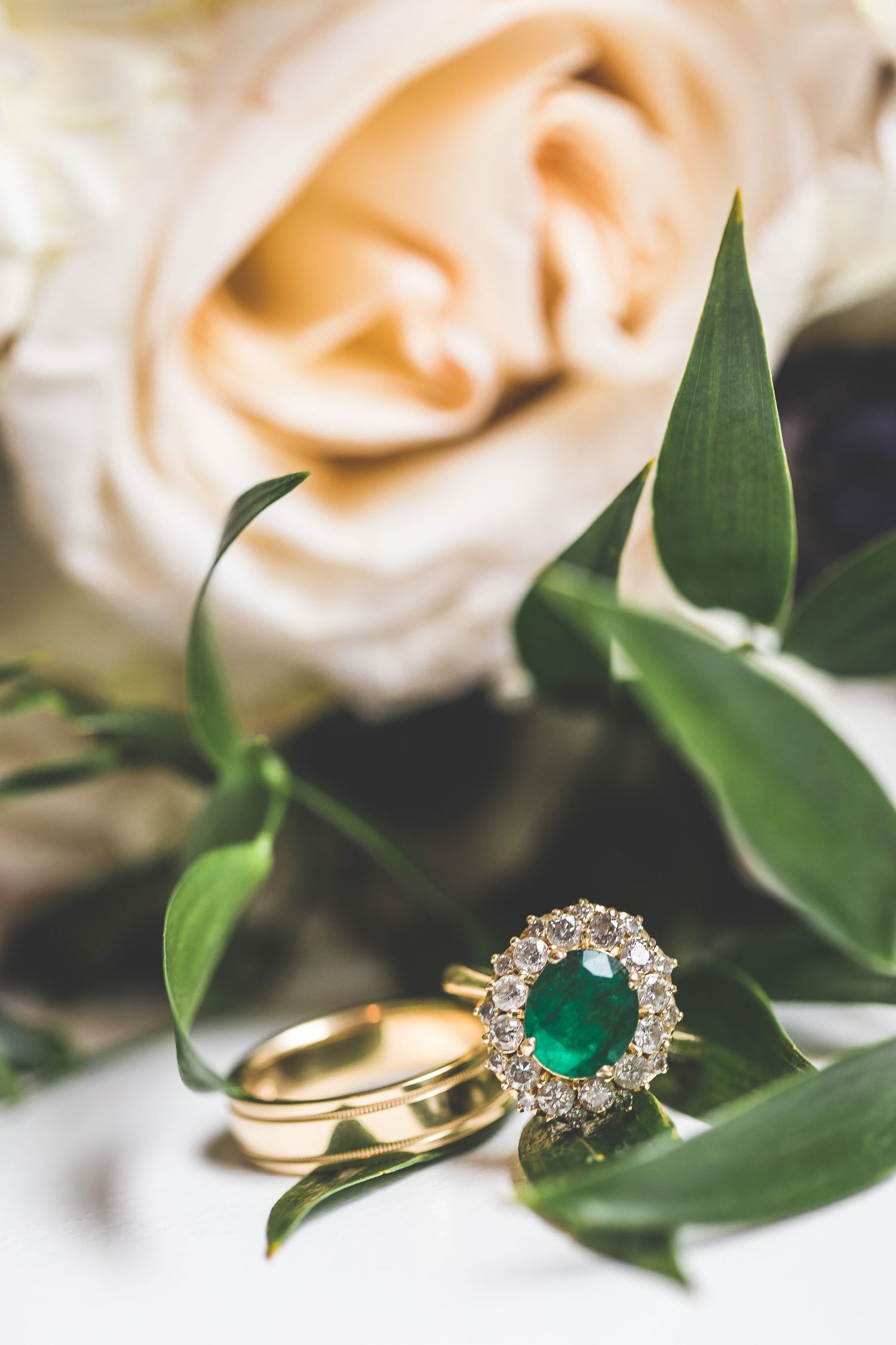 Wedding rings and emerald jewelry displayed among soft pink roses and green leaves.