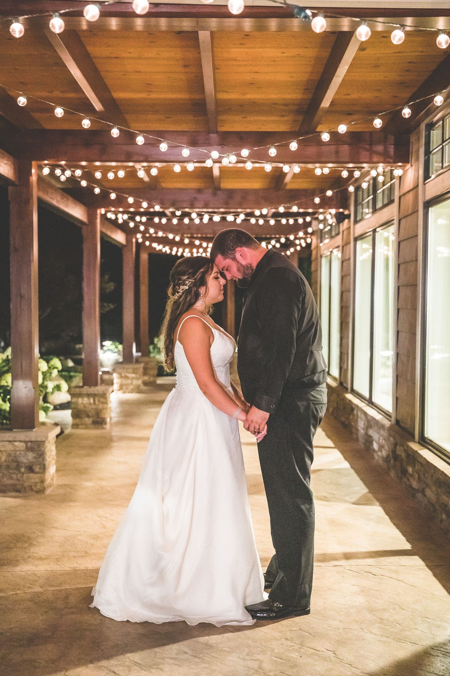 Wedding couple dancing under string lights in rustic covered porch setting.