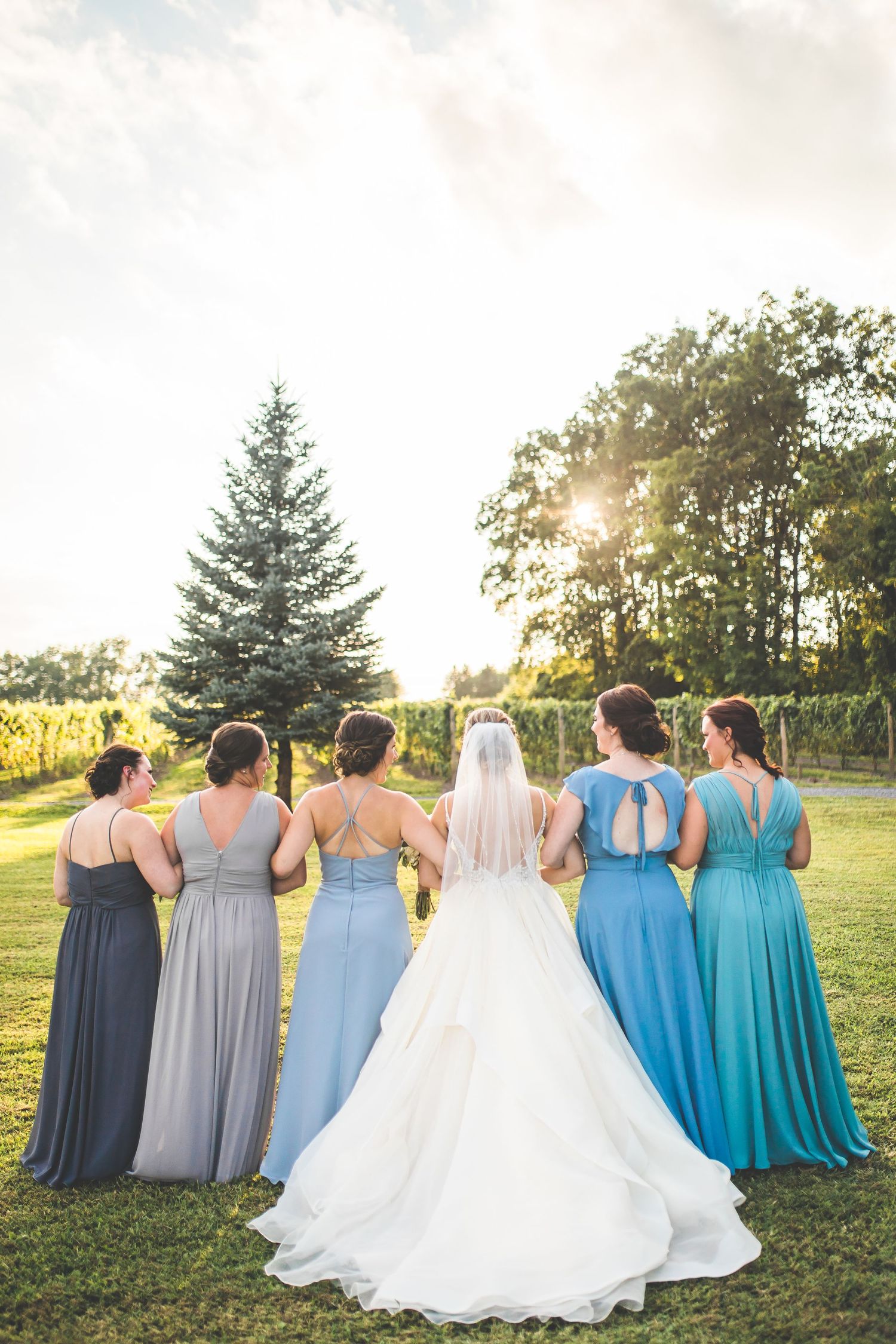 Bridesmaids in varying shades of blue dresses stand together with bride in flowing white gown.