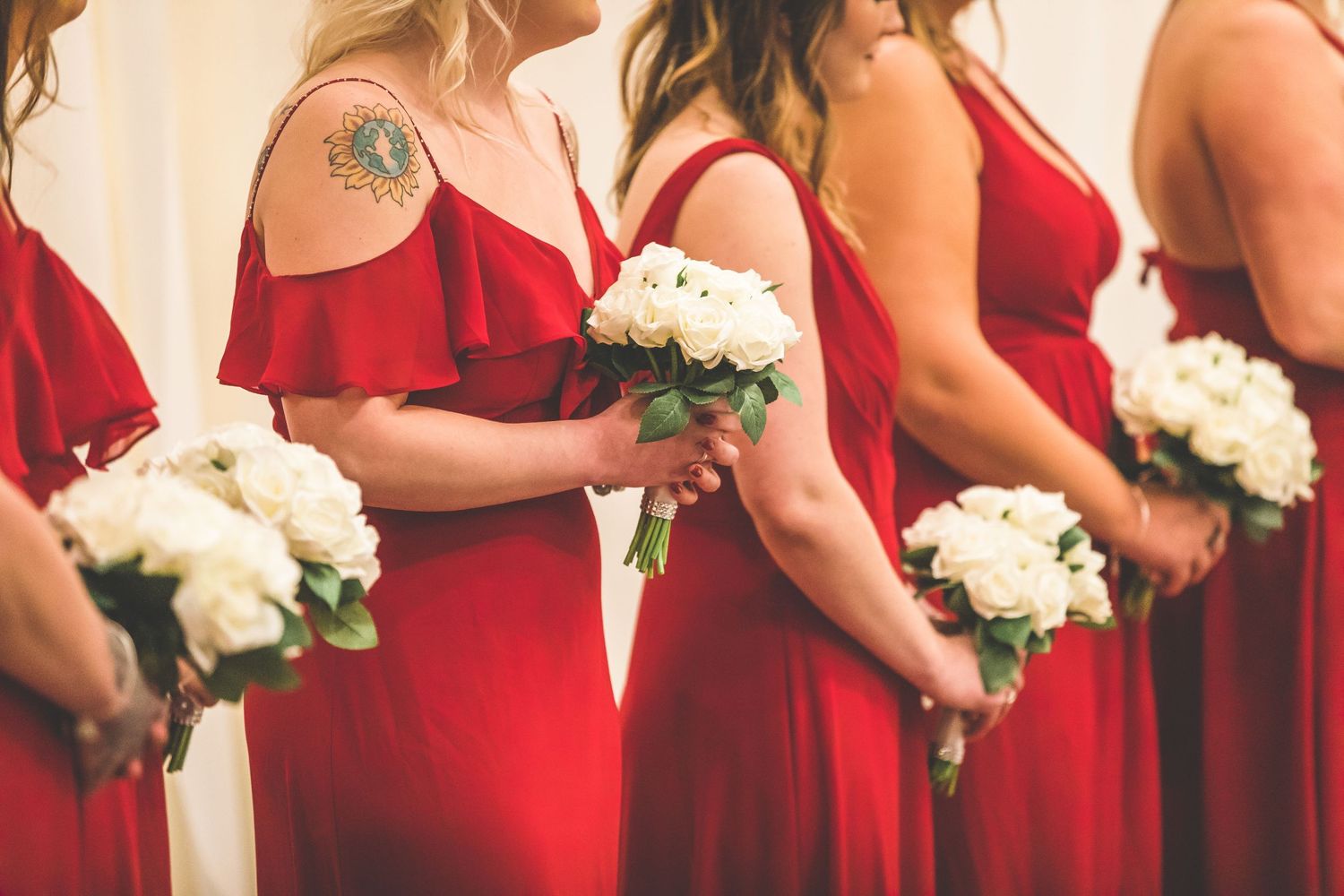 Bridesmaids in red dresses hold white bouquets while standing in line.