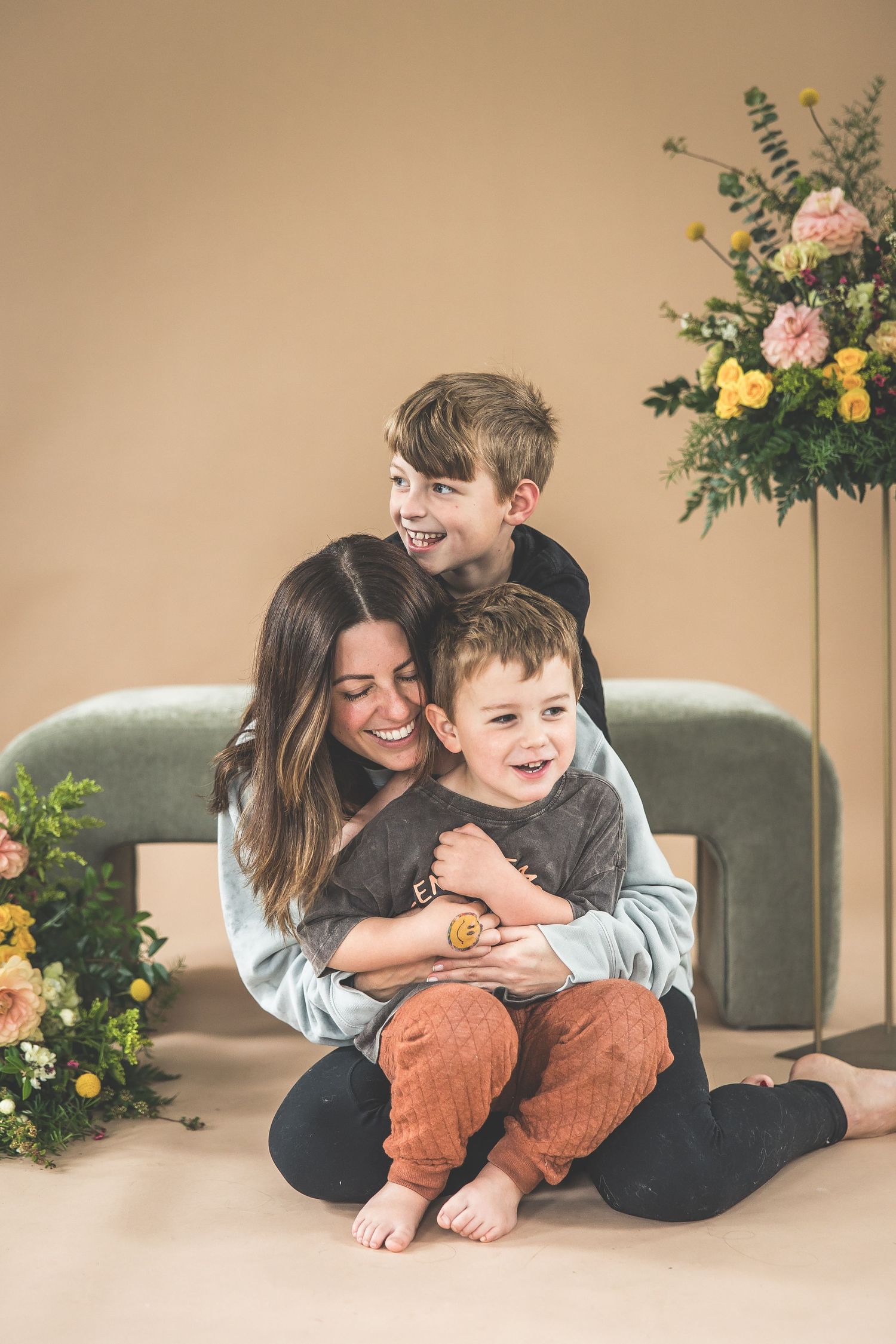 A smiling woman sits on the floor hugging two young boys in a warmly decorated studio with floral arrangements.