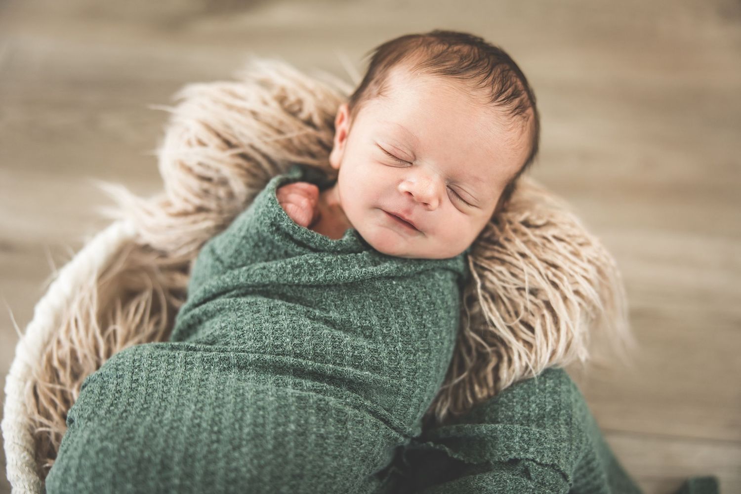A peaceful sleeping newborn wrapped in green knit blanket on soft fur backdrop.