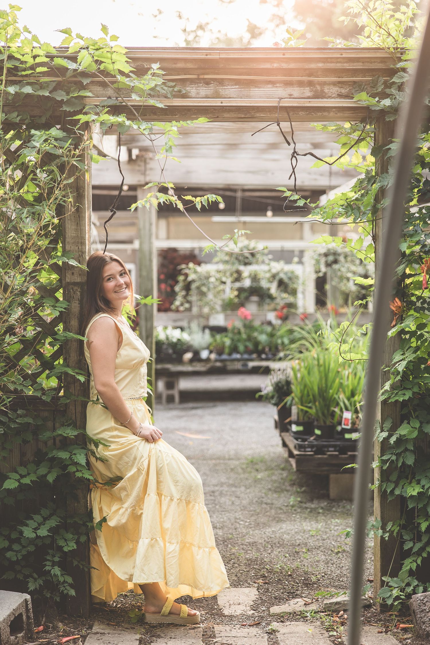 Woman in yellow sundress poses gracefully in a lush garden greenhouse setting.