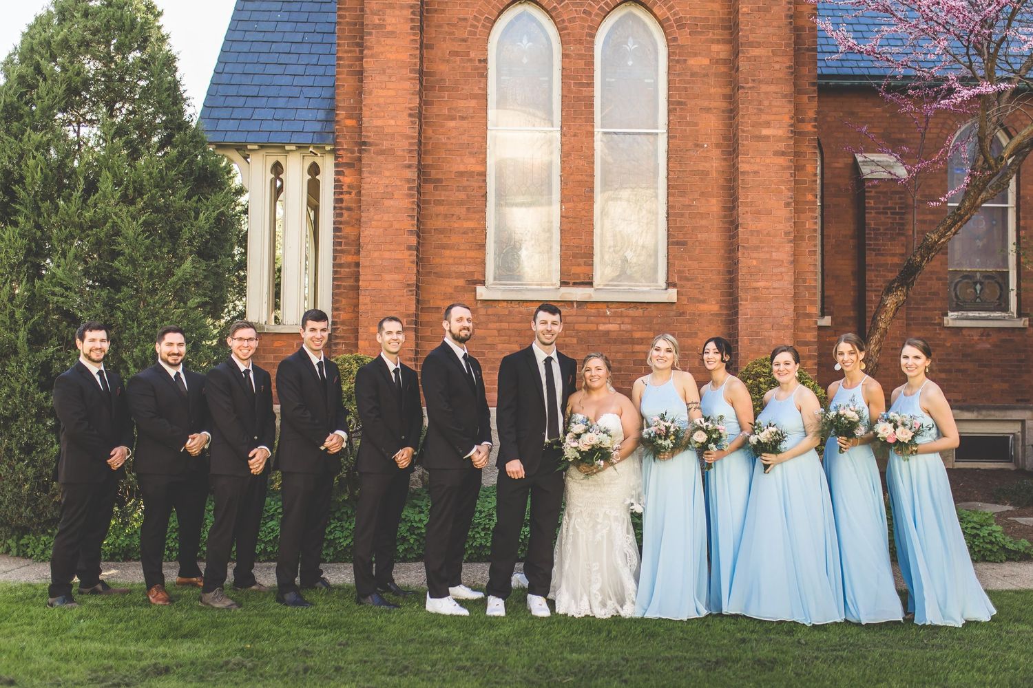 Large wedding party lined up outside red brick church with bridesmaids in light blue dresses.