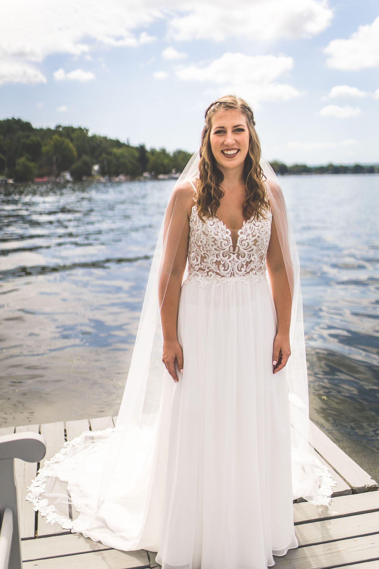 Bride in lace-detailed wedding gown stands on wooden dock overlooking lake.