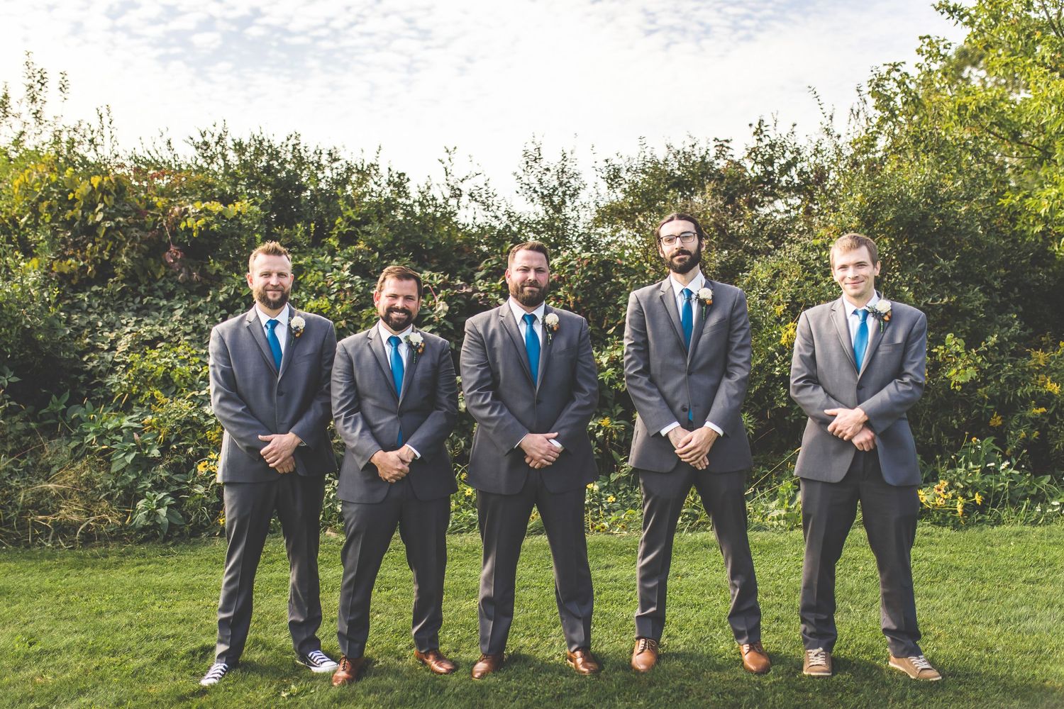 Groomsmen in matching suits pose casually on grassy hillside during wedding celebration.