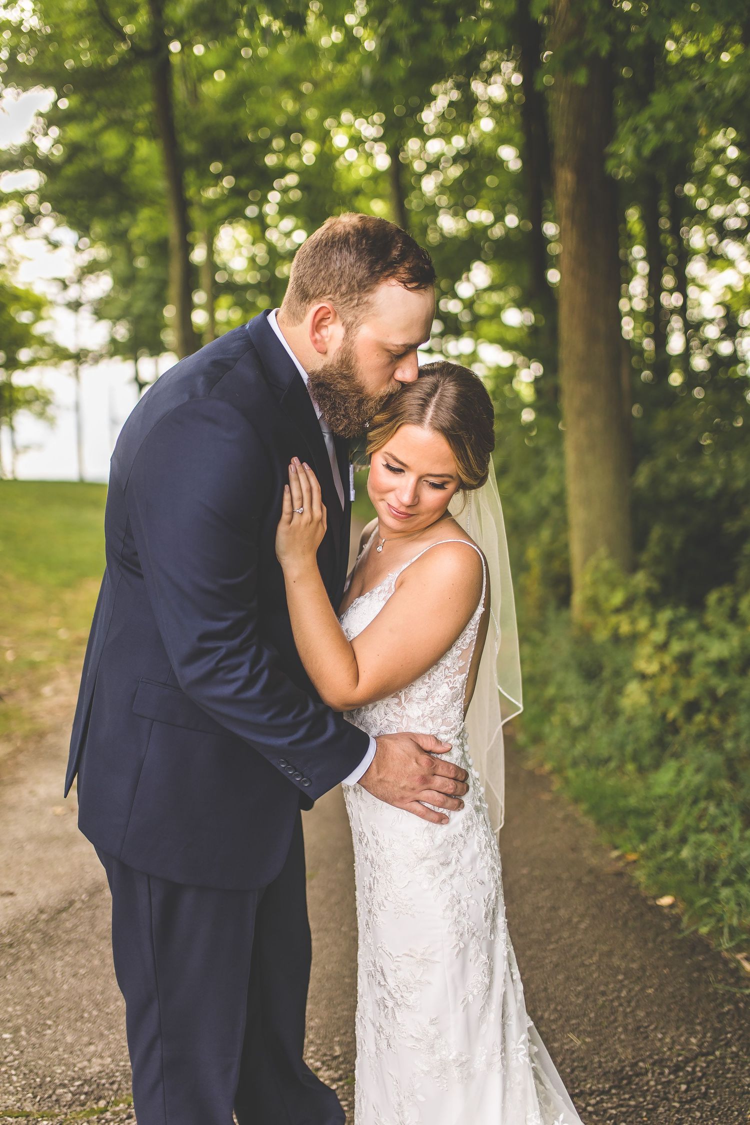 Bride in lace dress and groom in navy suit embrace on a wooded path during golden hour.