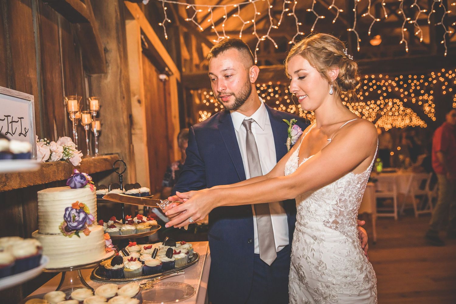 Couple cutting their wedding cake in rustic barn venue illuminated by string lights.