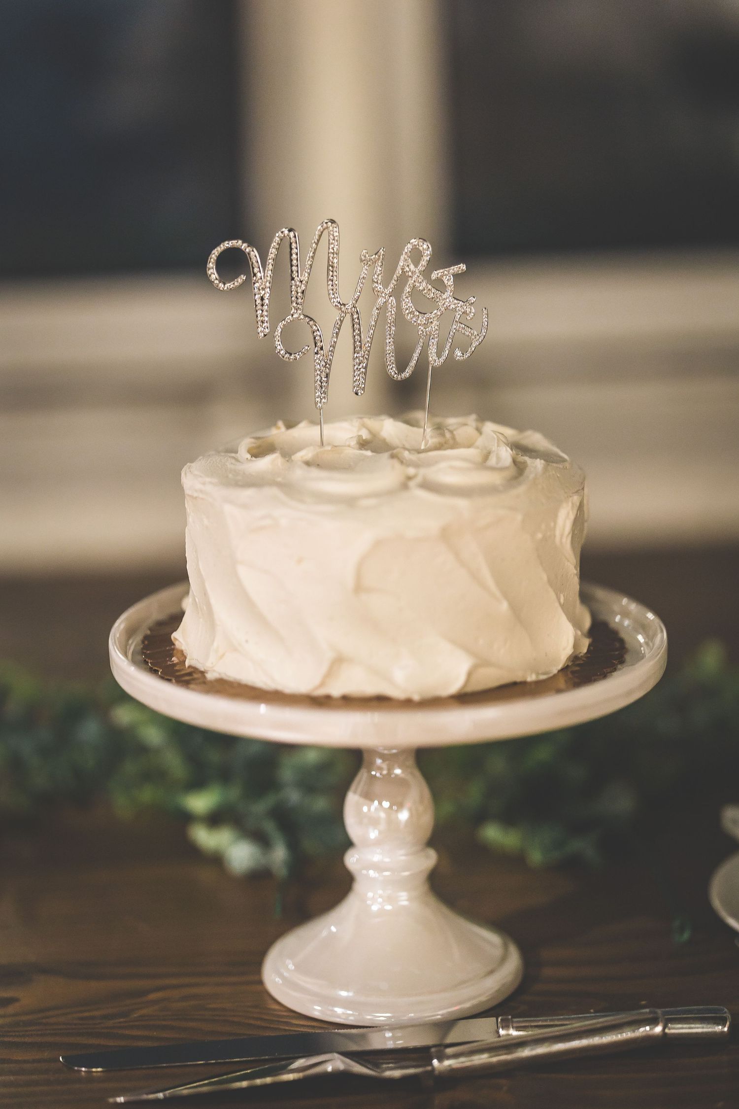 Simple white wedding cake displayed on an elegant cake stand with delicate topper decoration.
