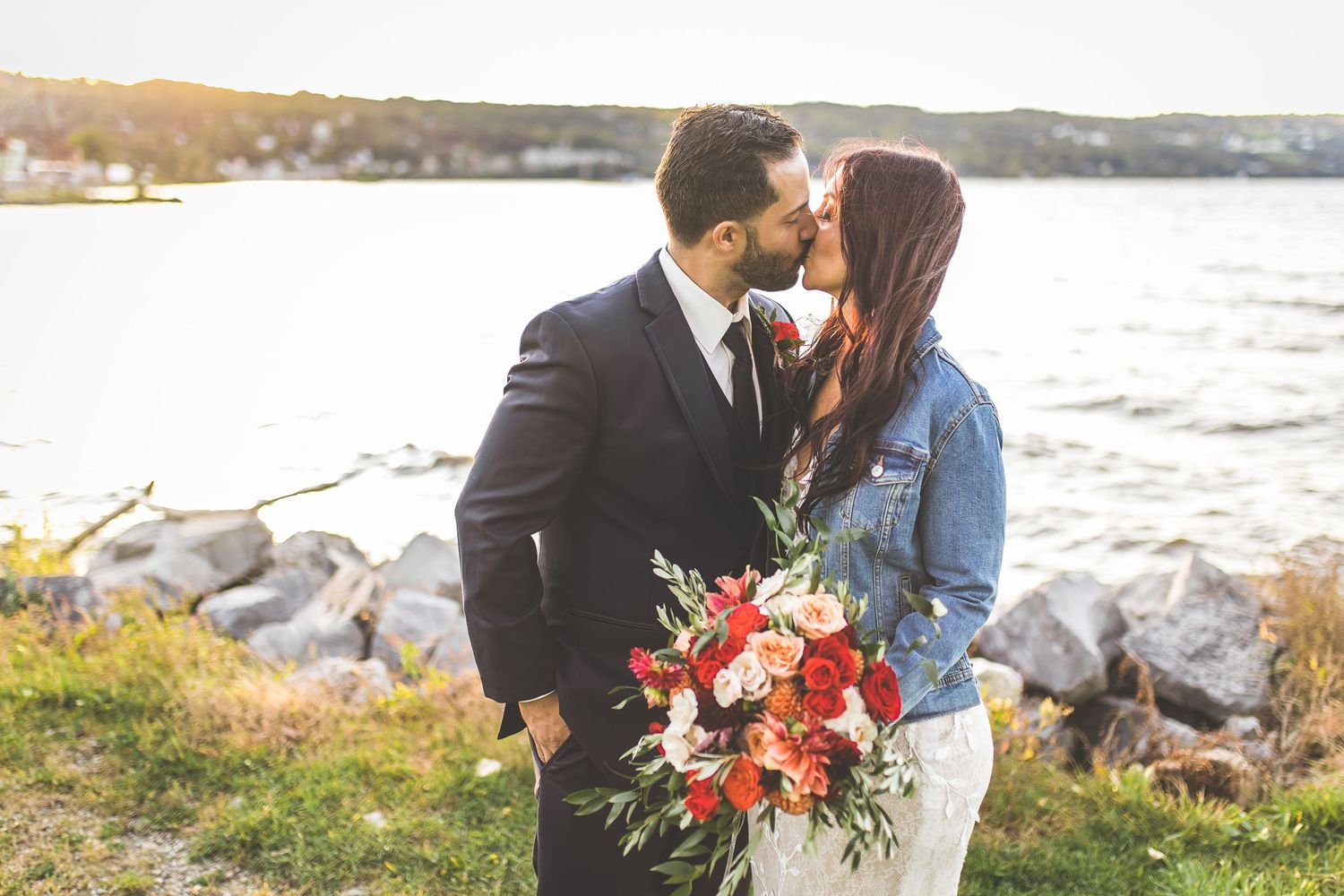 Couple in wedding attire embraces near waterfront with autumn colors.