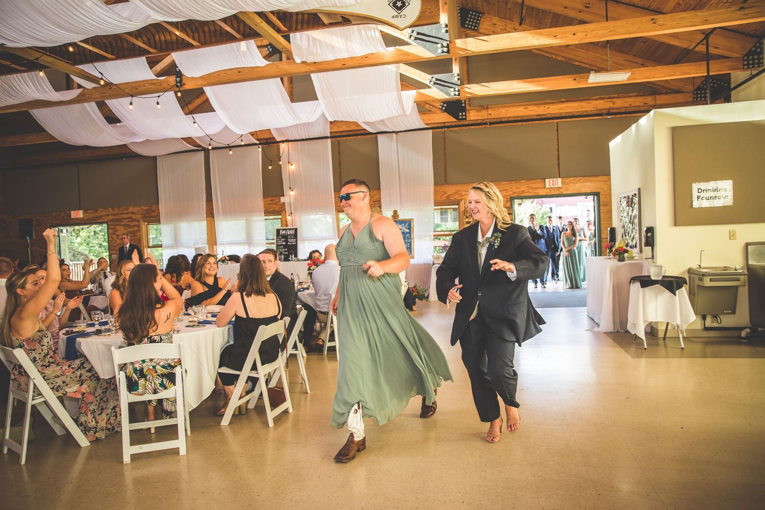 Wedding guests gather in a spacious reception hall with exposed wooden beams and white decor.