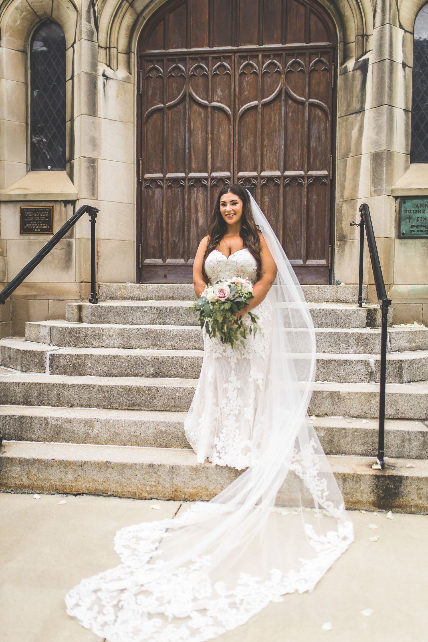 Bride in flowing lace gown stands on stone church steps with dramatic cathedral veil and bouquet.