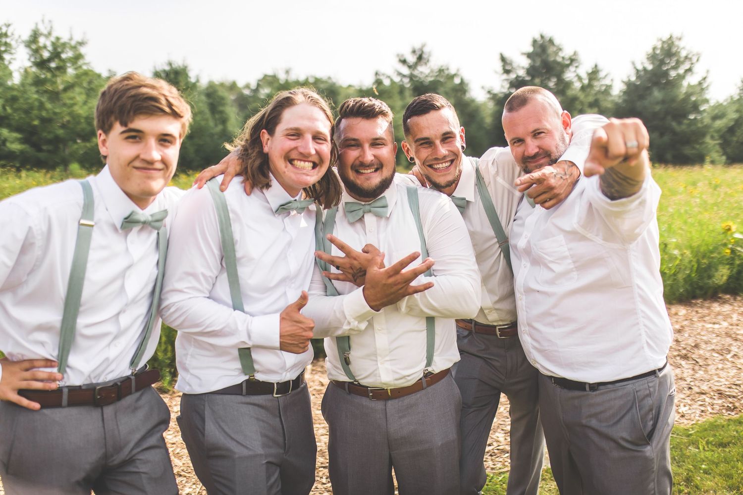 Groomsmen in white shirts and suspenders pose together laughing outdoors.