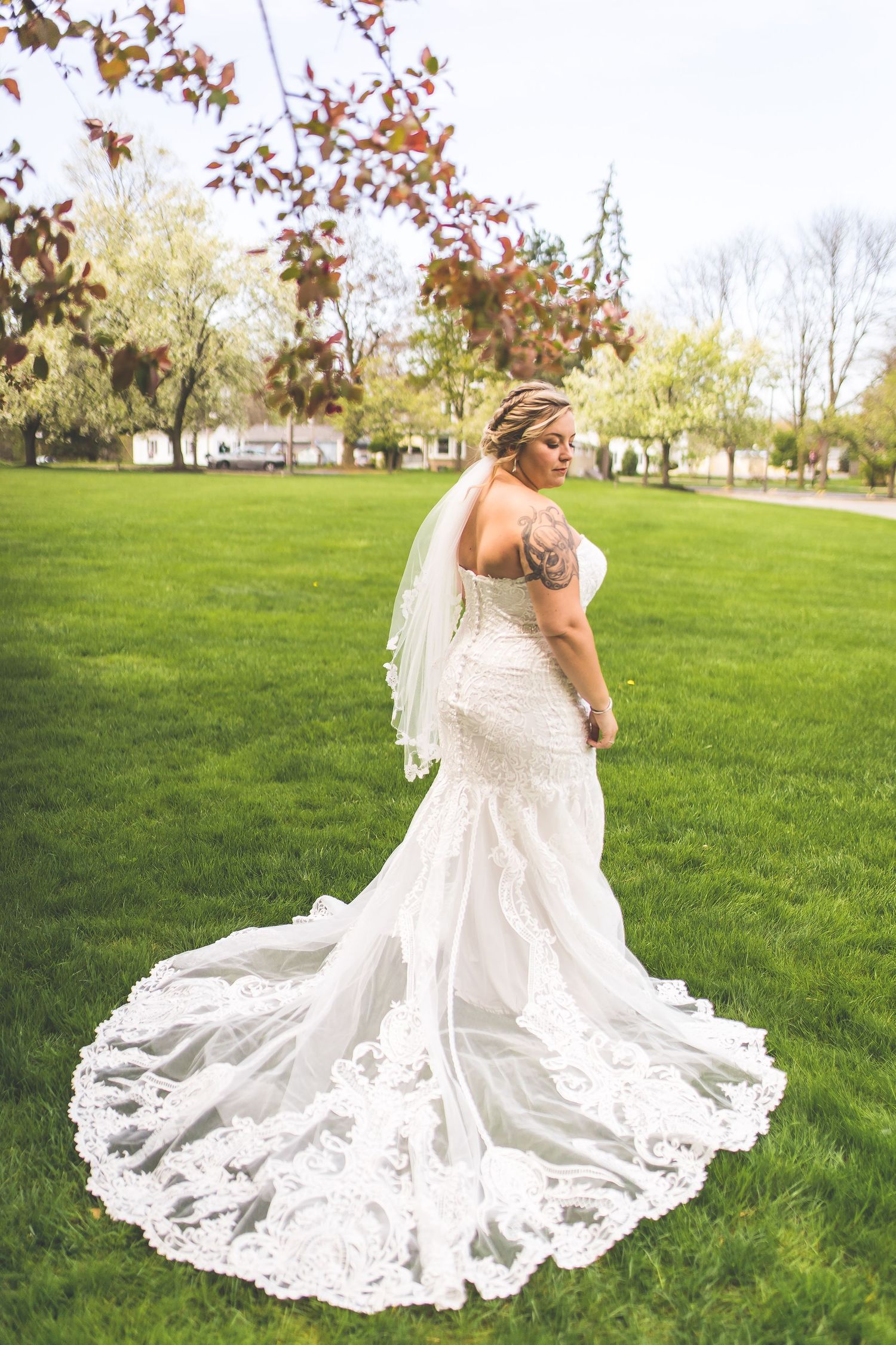 A wedding dress with intricate lace train flows across green grass during an outdoor bridal shoot.