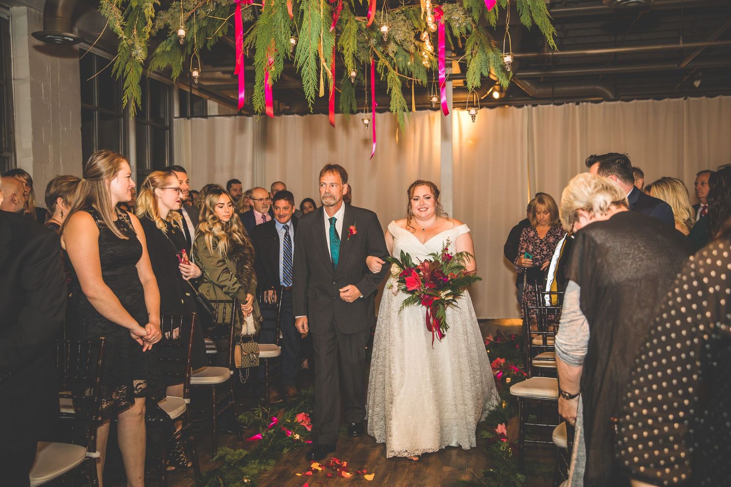 Large wedding reception crowd gathered in an industrial-style venue with hanging greenery and lights.