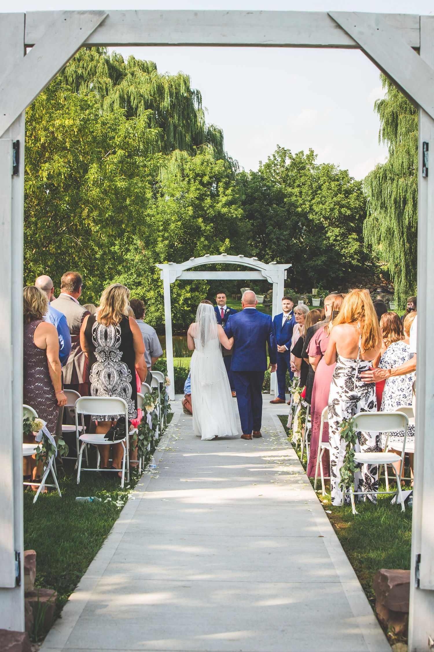 Wedding guests seated outdoors under a white pergola with willow trees and greenery in the background.