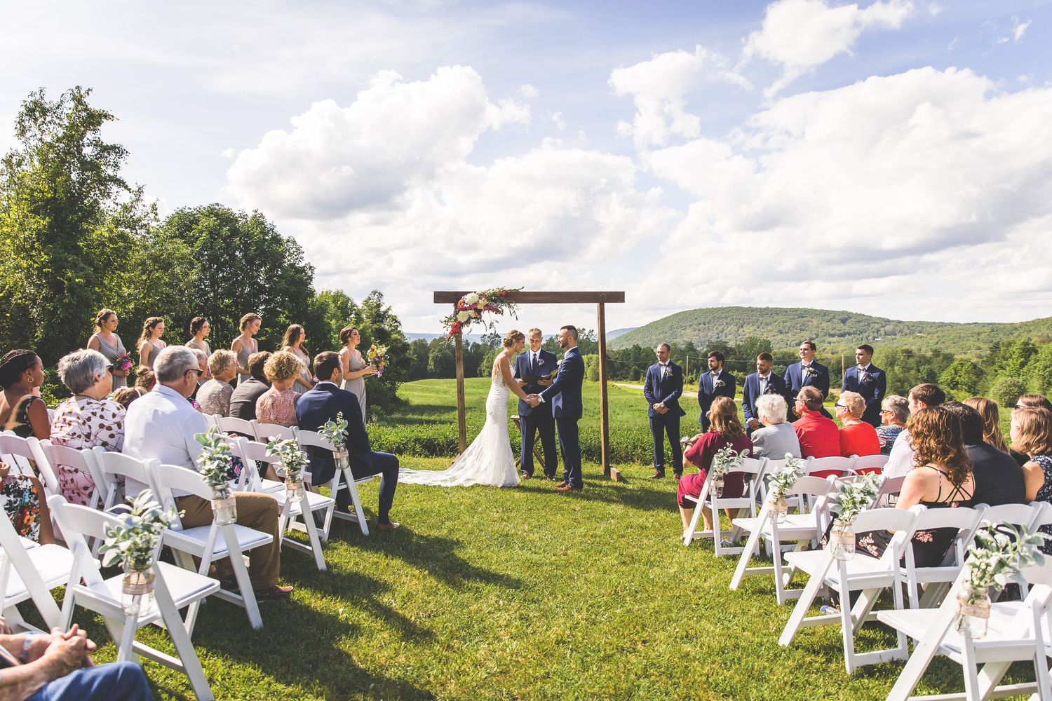 Outdoor wedding ceremony setup with white chairs on a grassy hillside under cloudy skies.