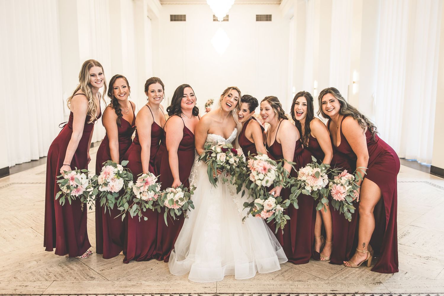Bridal party in burgundy dresses poses together in elegant white venue.