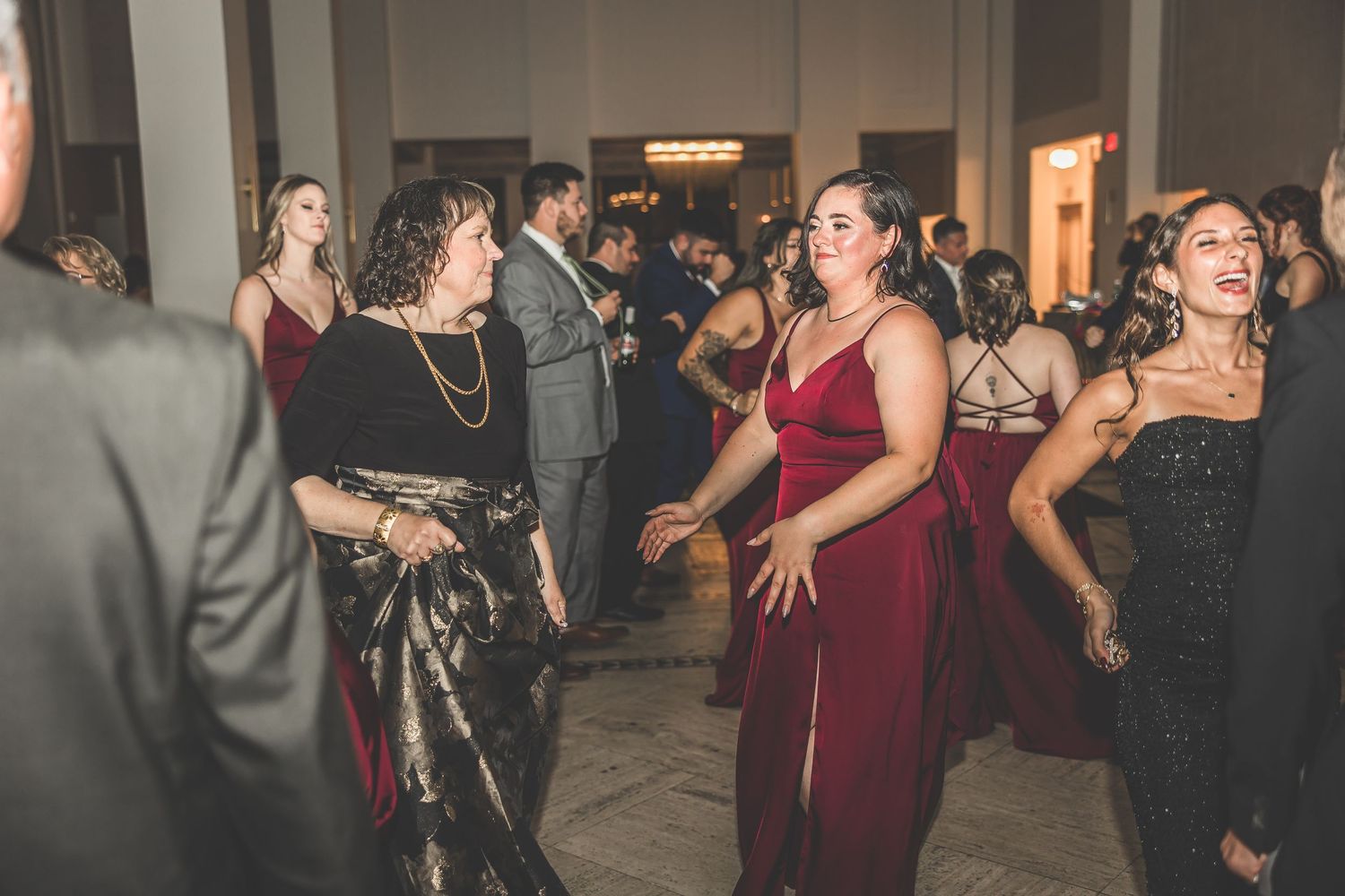 Wedding guests dance in formal attire under decorative lighting at an indoor venue.