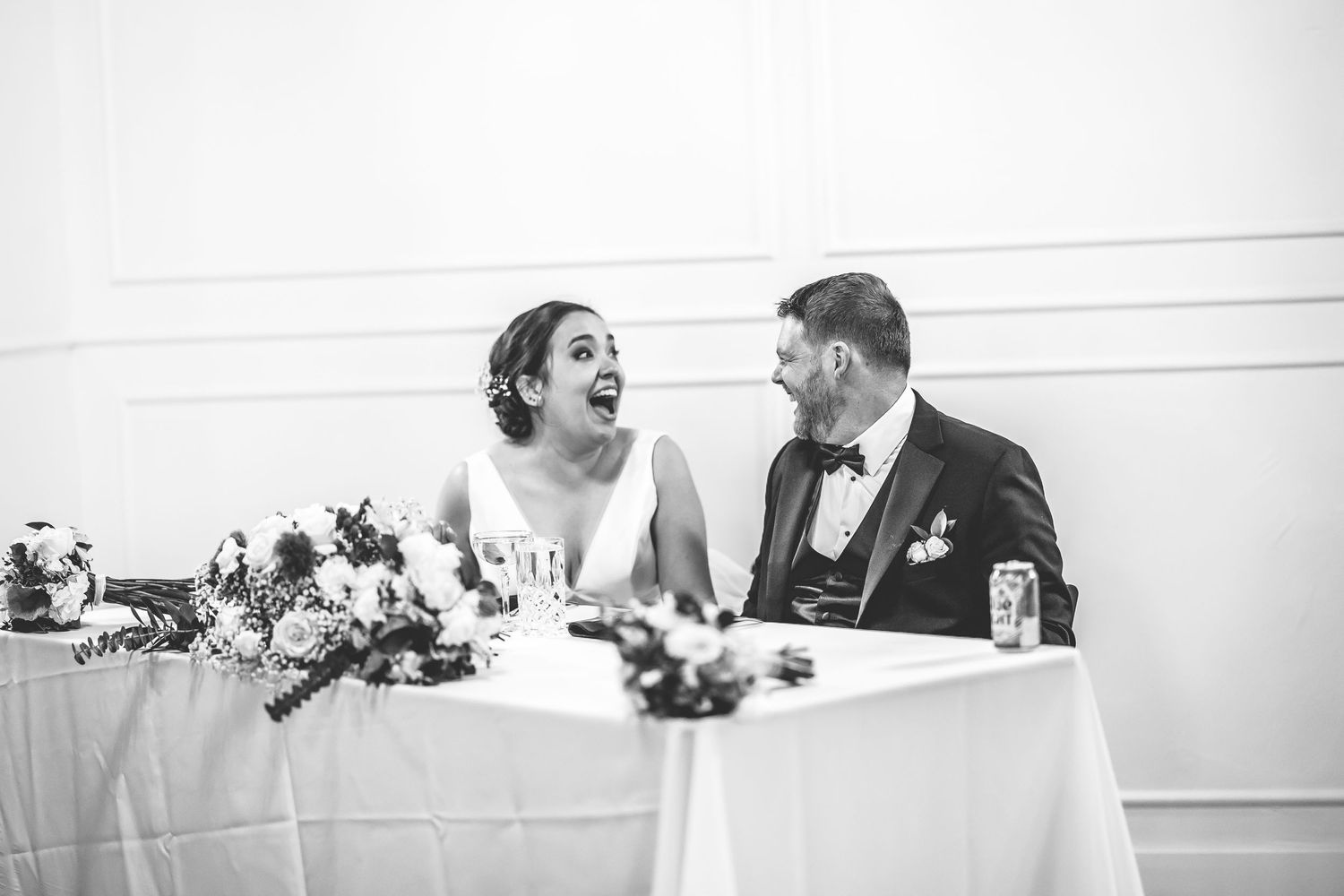 A candid black and white photo of a couple laughing at their reception table.