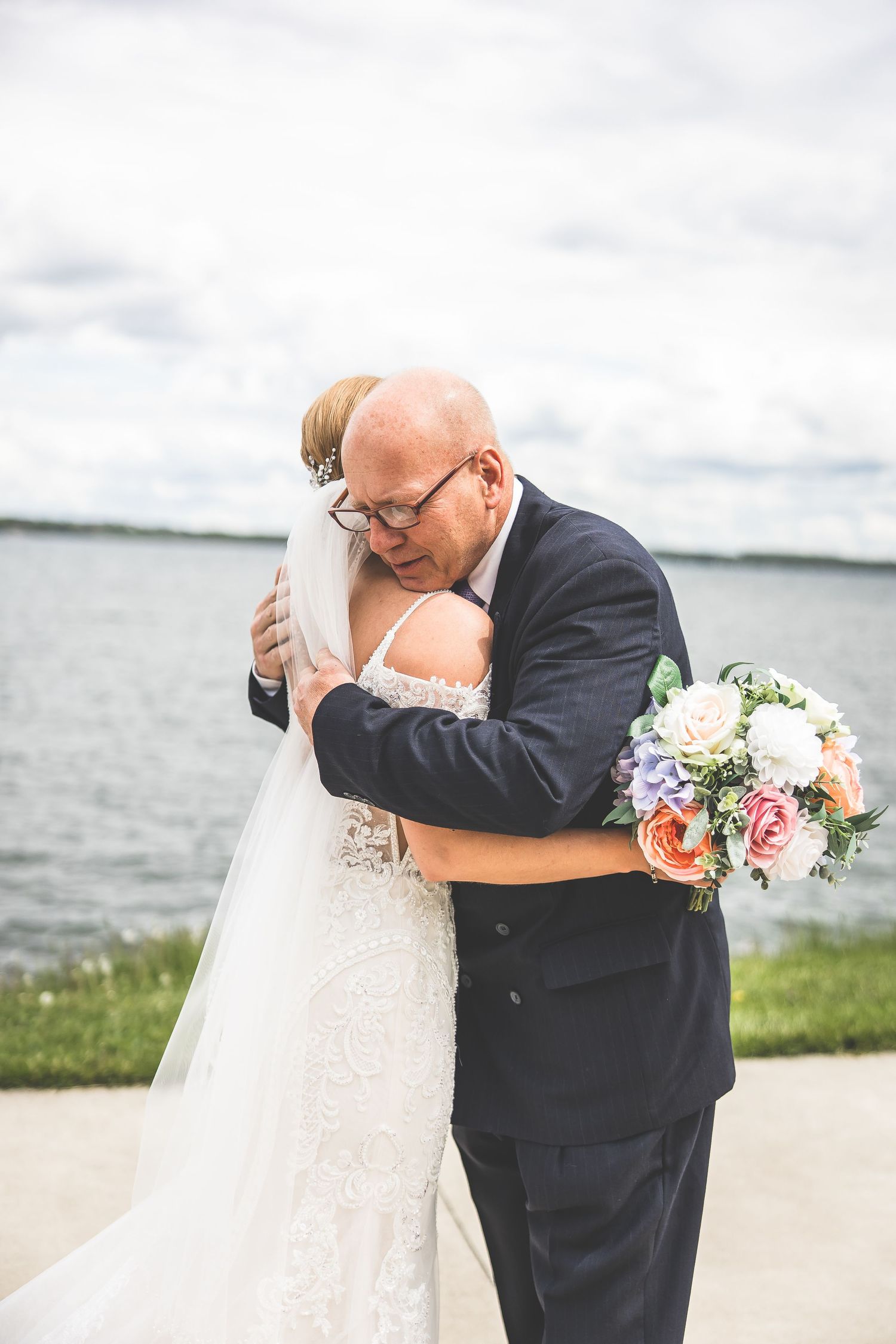 Emotional embrace during waterfront wedding ceremony with bouquet of pink and white flowers.