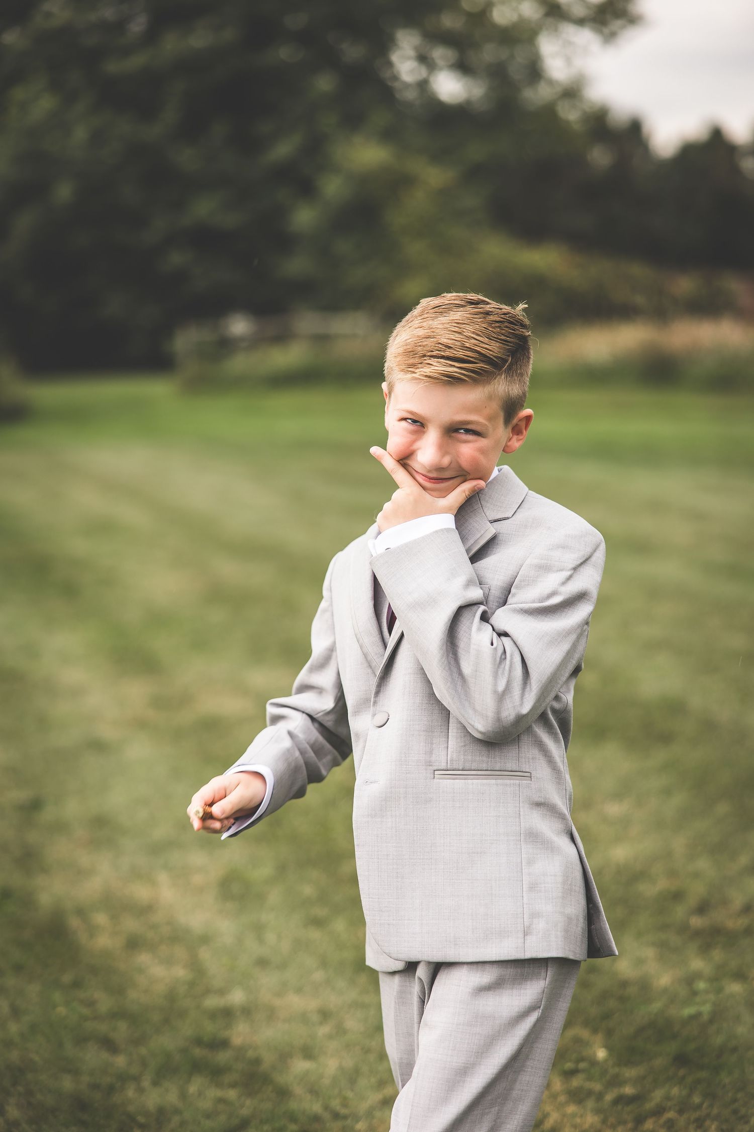 Young ring bearer in a light gray suit smiles playfully in an outdoor garden setting.