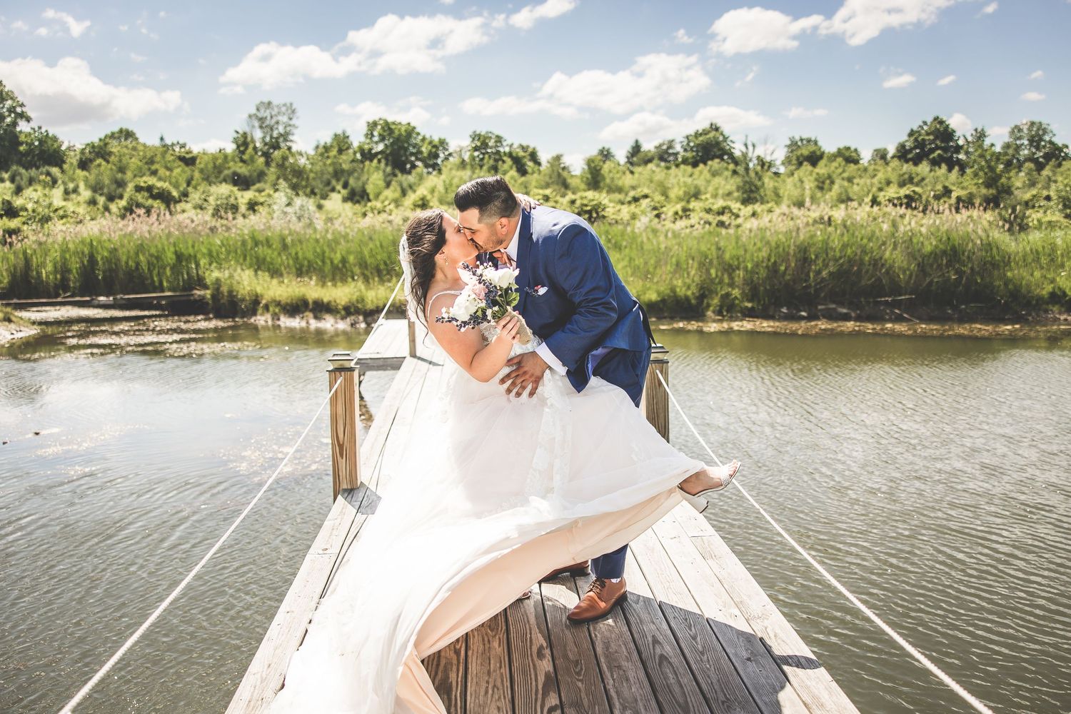 Wedding couple sharing romantic moments on a lakeside wooden dock under blue skies.