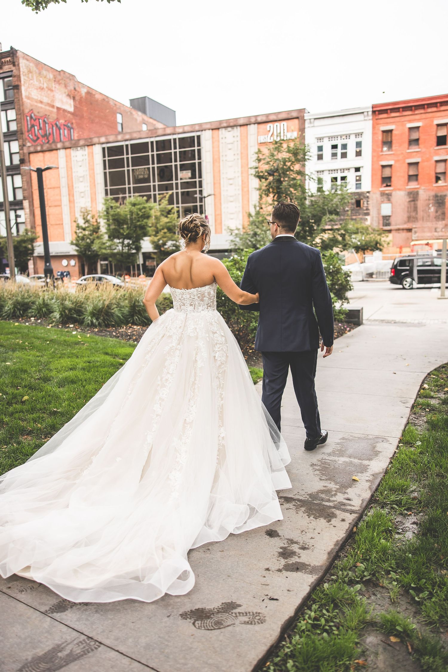 Wedding couple walking away from camera on urban pathway, bride's dress trailing behind.