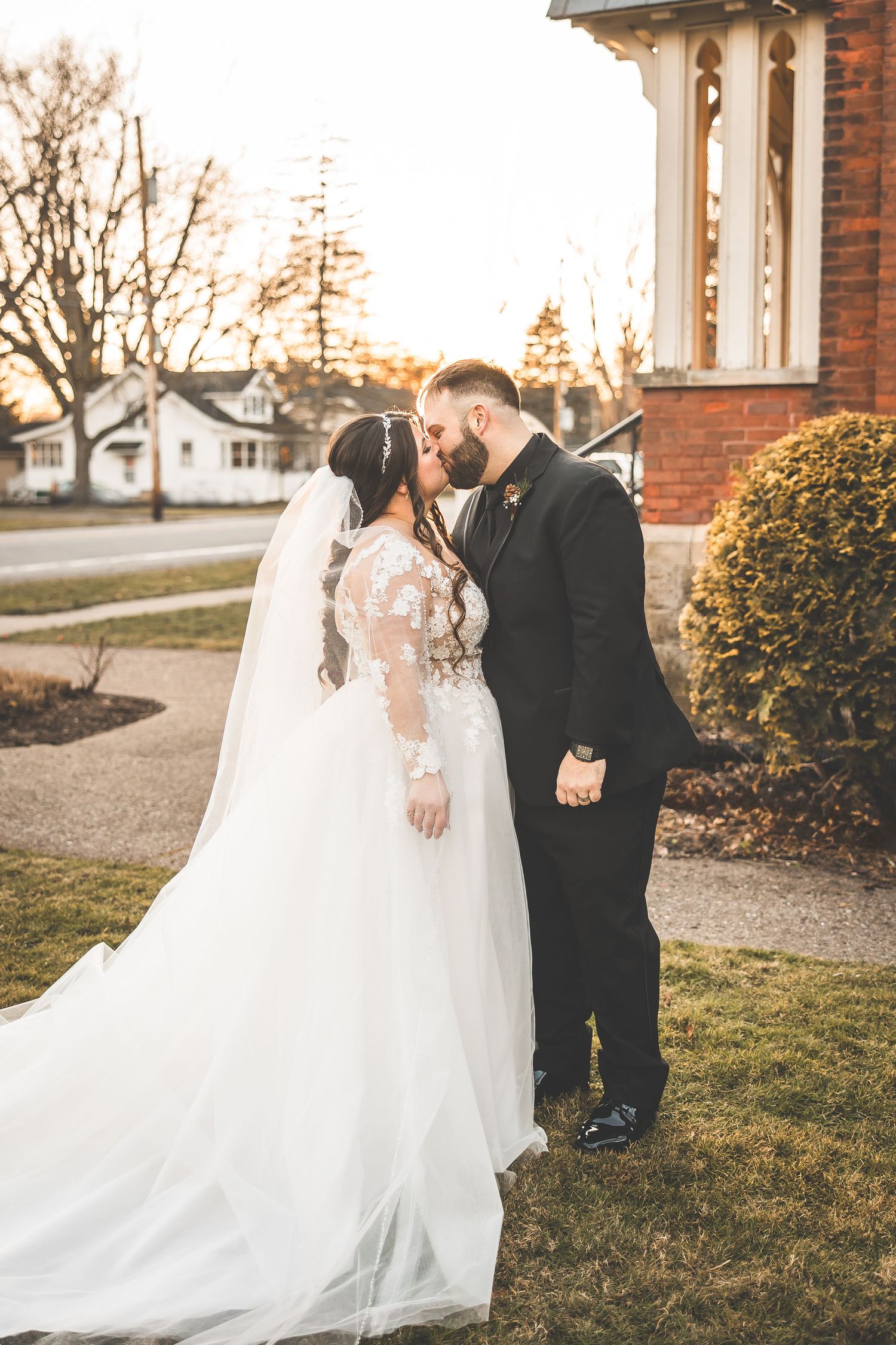 Bride in flowing white gown and groom in black suit pose at sunset near brick church.