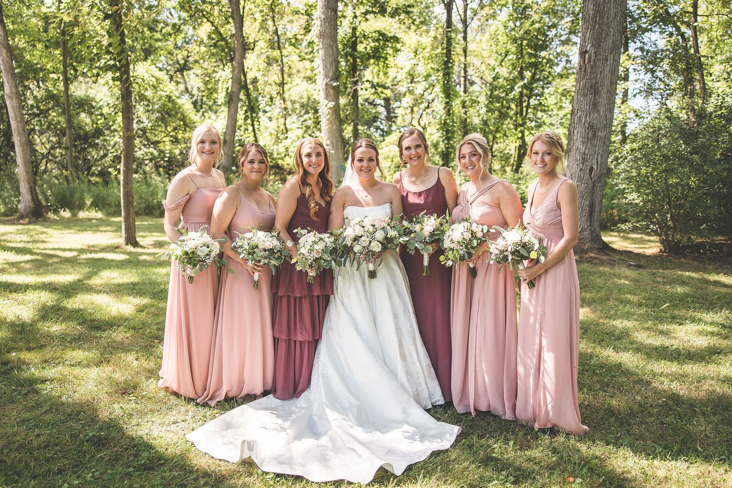 Bridal party in pink and burgundy dresses poses with bride in white gown outdoors.