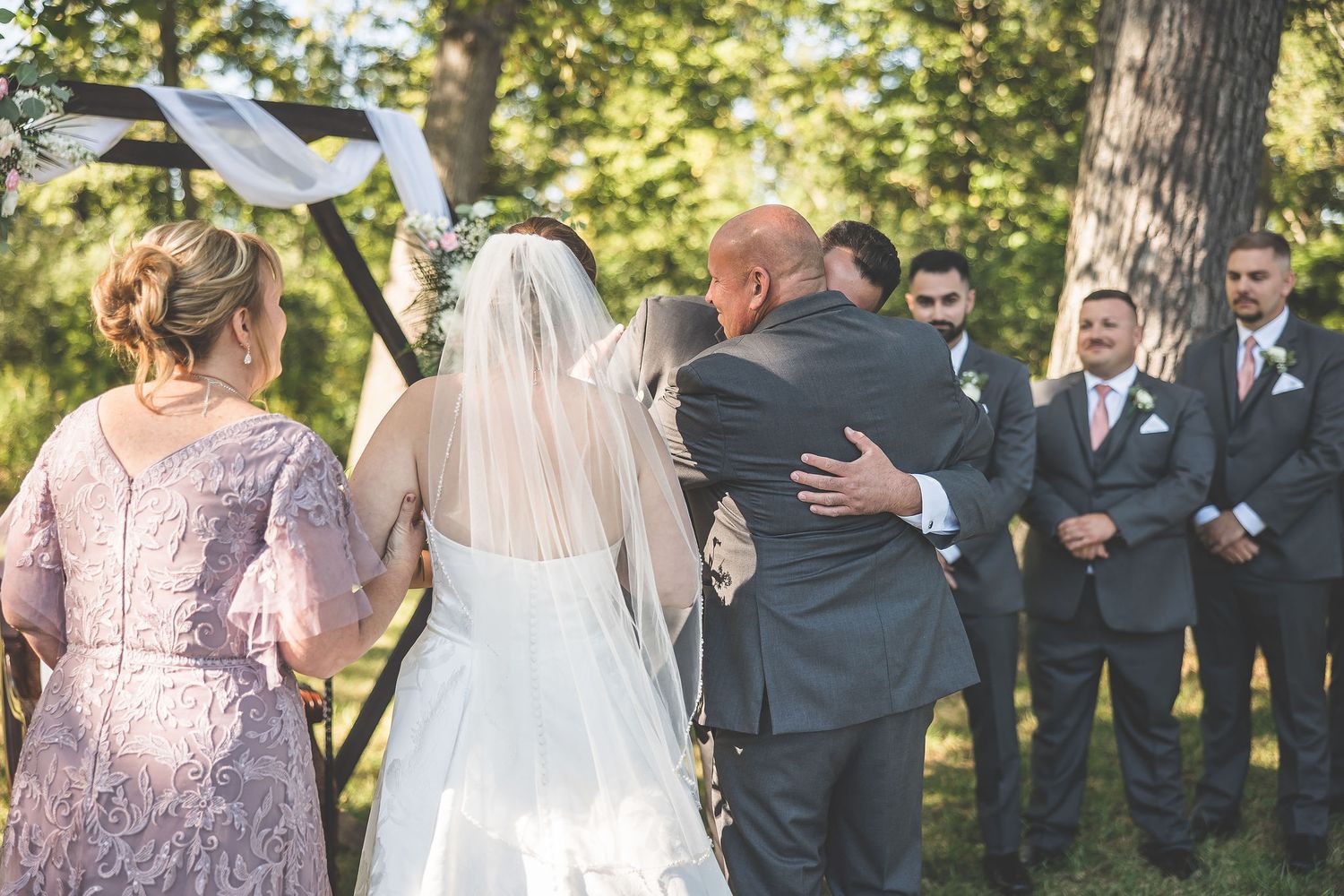 Wedding ceremony under trees with white fabric draped arch and floral arrangements.