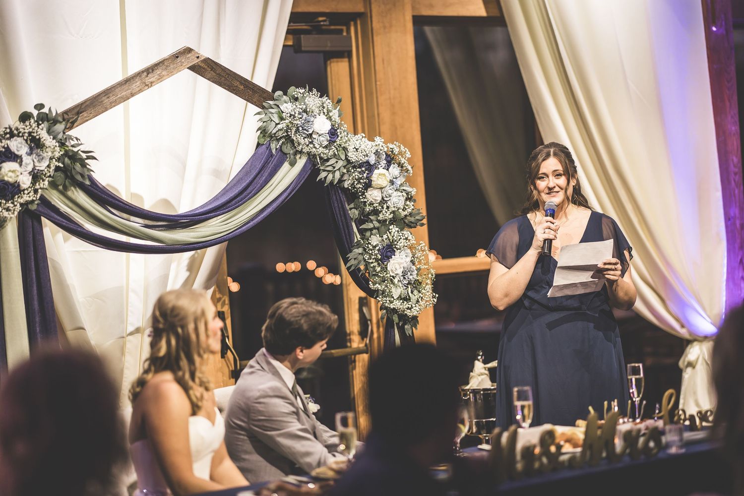 Wedding speech being delivered at reception with decorated arch and white floral arrangements.