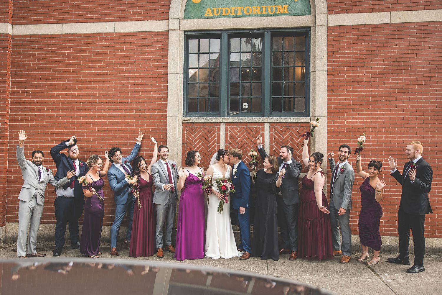 Large wedding party group photo outside a traditional red brick building entrance.
