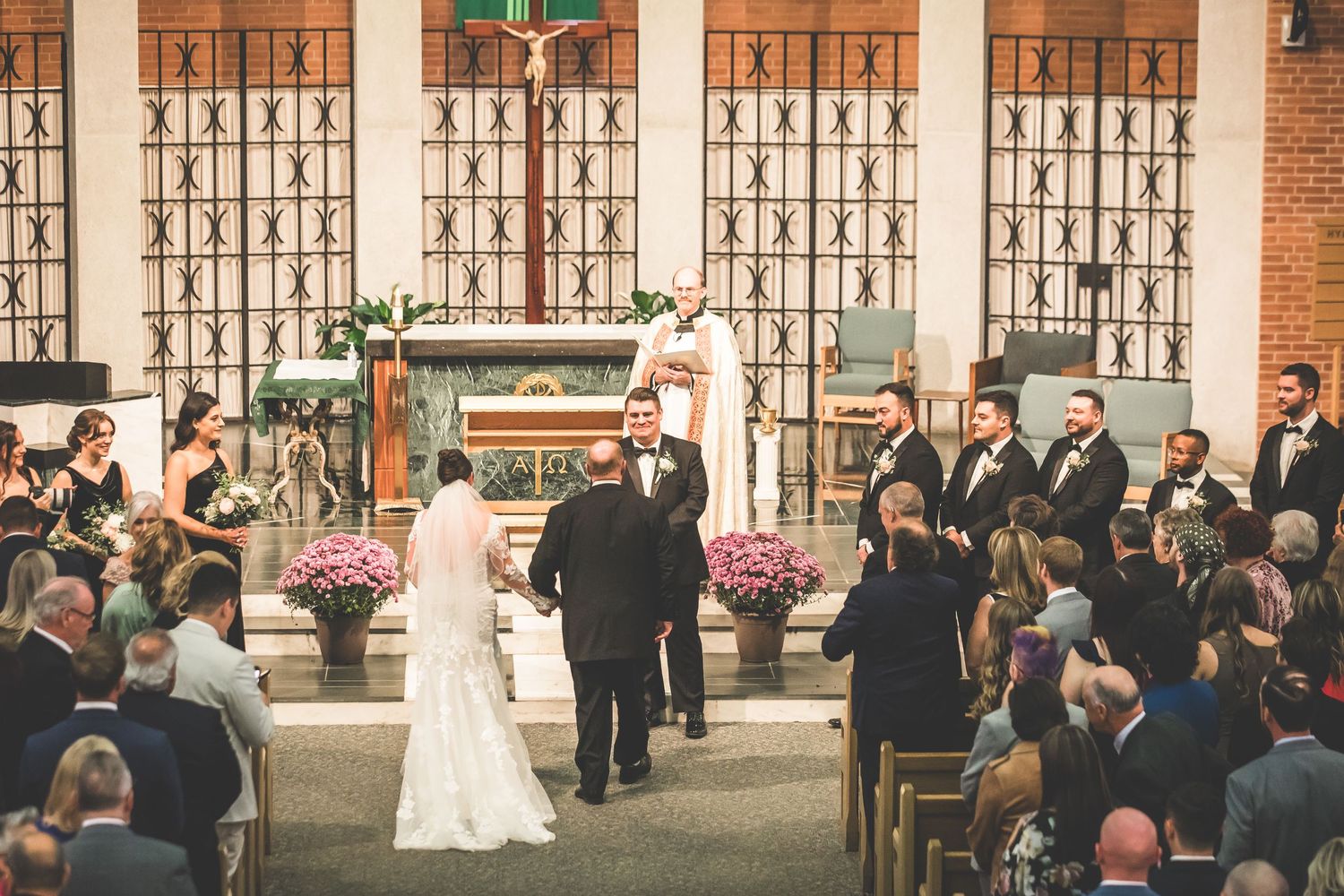 Wedding ceremony at church altar with guests seated in pews and pink floral arrangements.