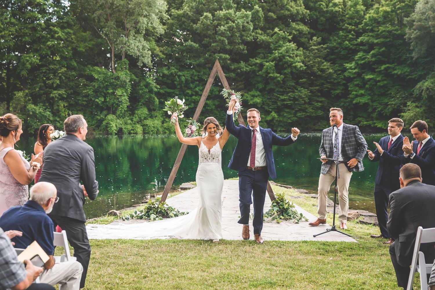 Wedding guests seated in white chairs during outdoor ceremony with wooden arch.