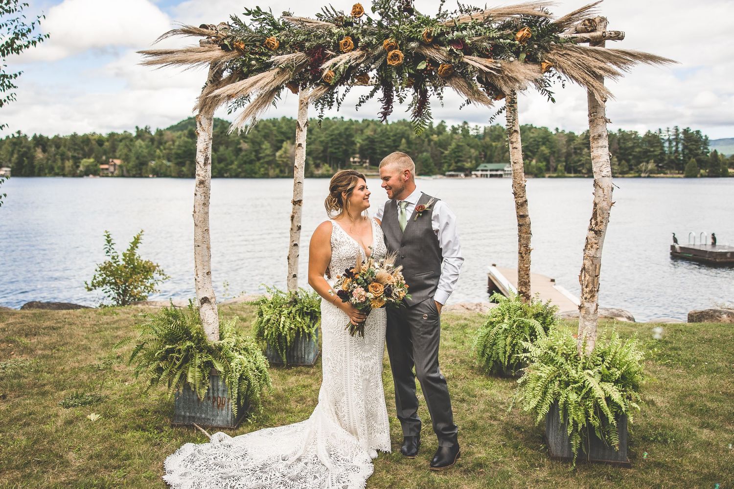 Lakeside wedding ceremony setup with decorated birch branch arch and natural landscaping.