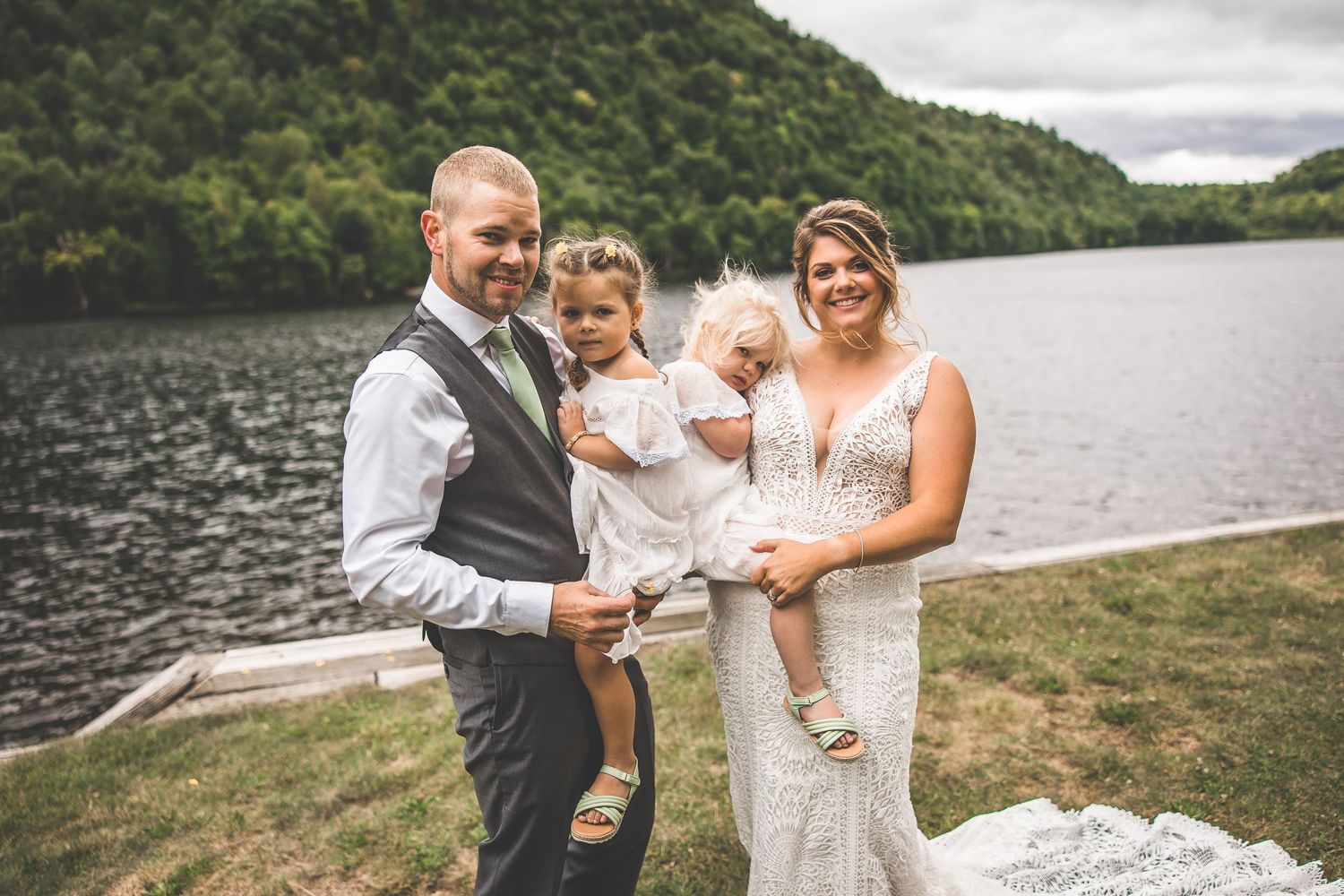 Wedding couple and child stand together by a scenic mountain lake on an overcast day.