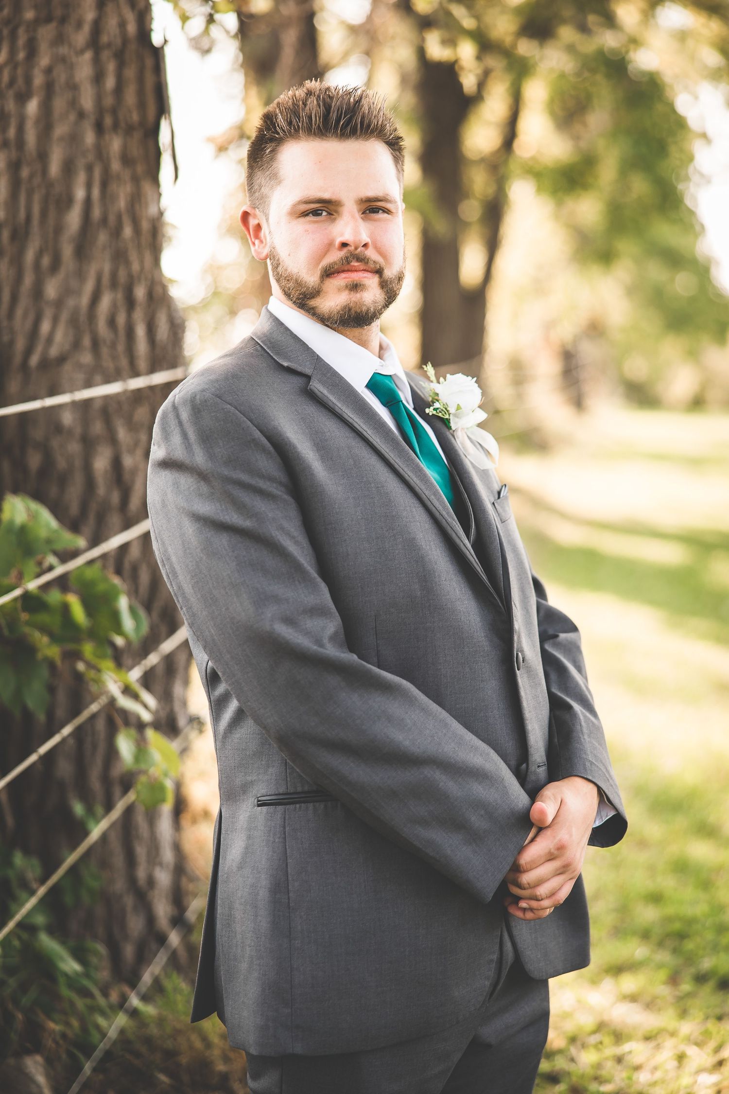 Groom in gray suit with teal tie poses against tree trunk in outdoor setting.