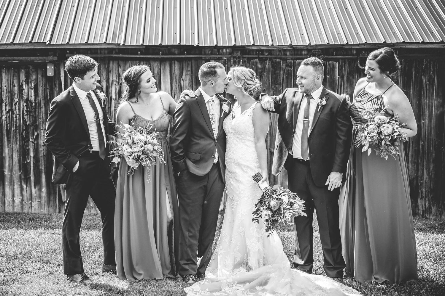 Wedding party members stand in formal line against weathered wooden barn wall.