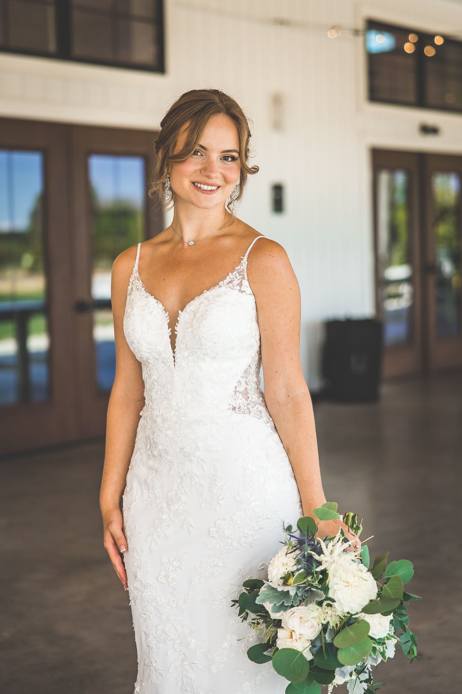 Bride in stunning white lace gown holding fresh floral bouquet in modern venue.