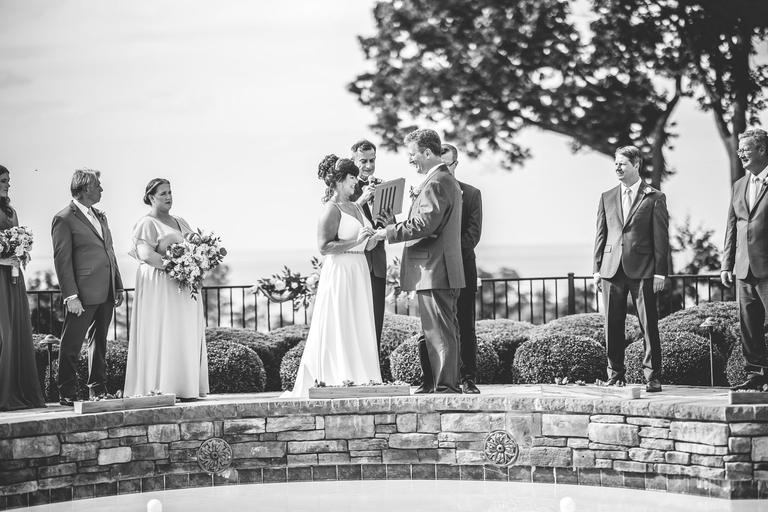 Black and white photo of wedding celebration on elegant stone terrace with trees in background.