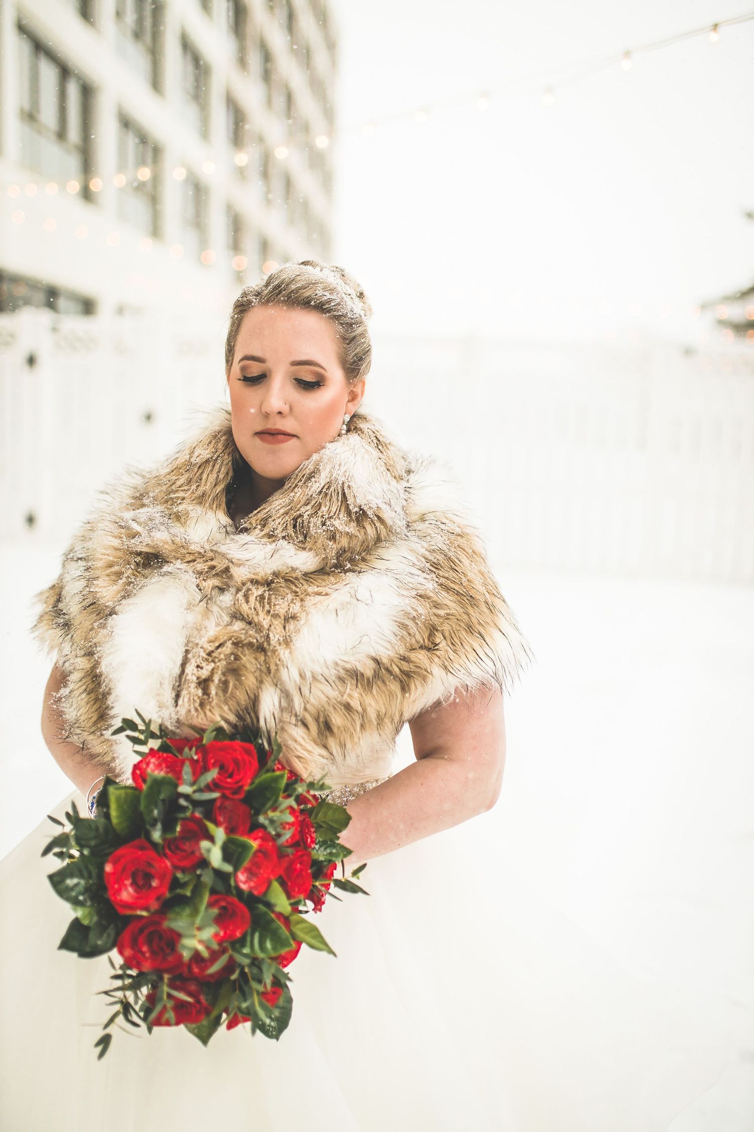 Winter bride wearing a fur stole holds a vibrant red bouquet against snowy backdrop.