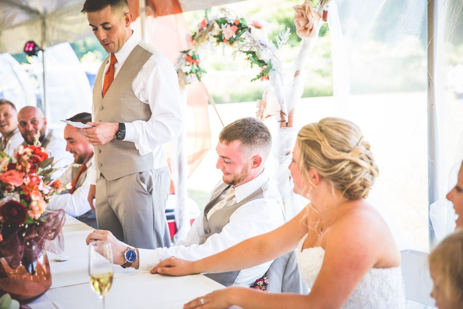 Wedding party members celebrating during an outdoor reception under white tent canopy.