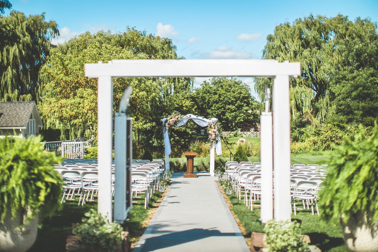 Outdoor wedding ceremony setup with white chairs and arbor along a garden path.
