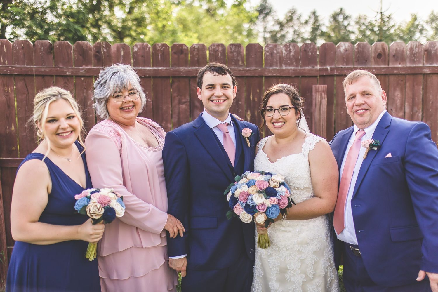 Wedding party members pose together with bouquets against a wooden fence in a garden setting.