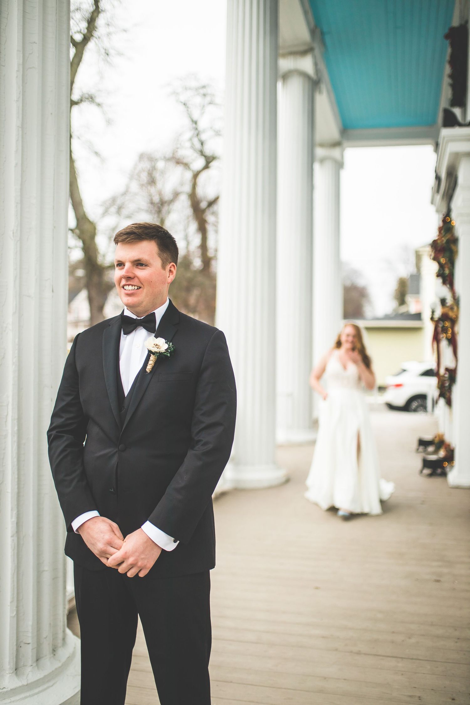 A groom in a black tuxedo standing on a columned porch waiting for the first look.