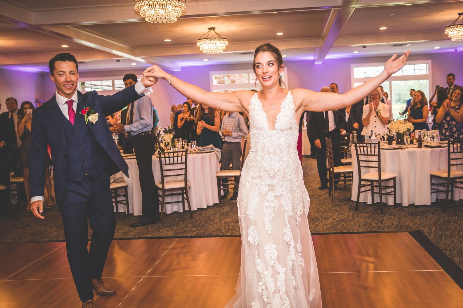 Newlyweds sharing their first dance in an elegant ballroom with purple ambient lighting and chandeliers.