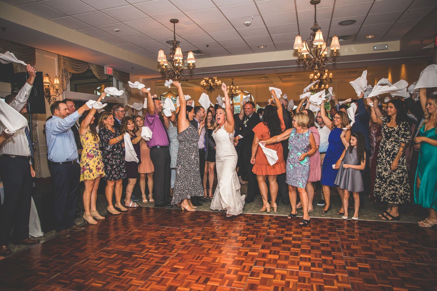 Wedding guests celebrate on a wooden dance floor under elegant chandeliers at an indoor reception venue.