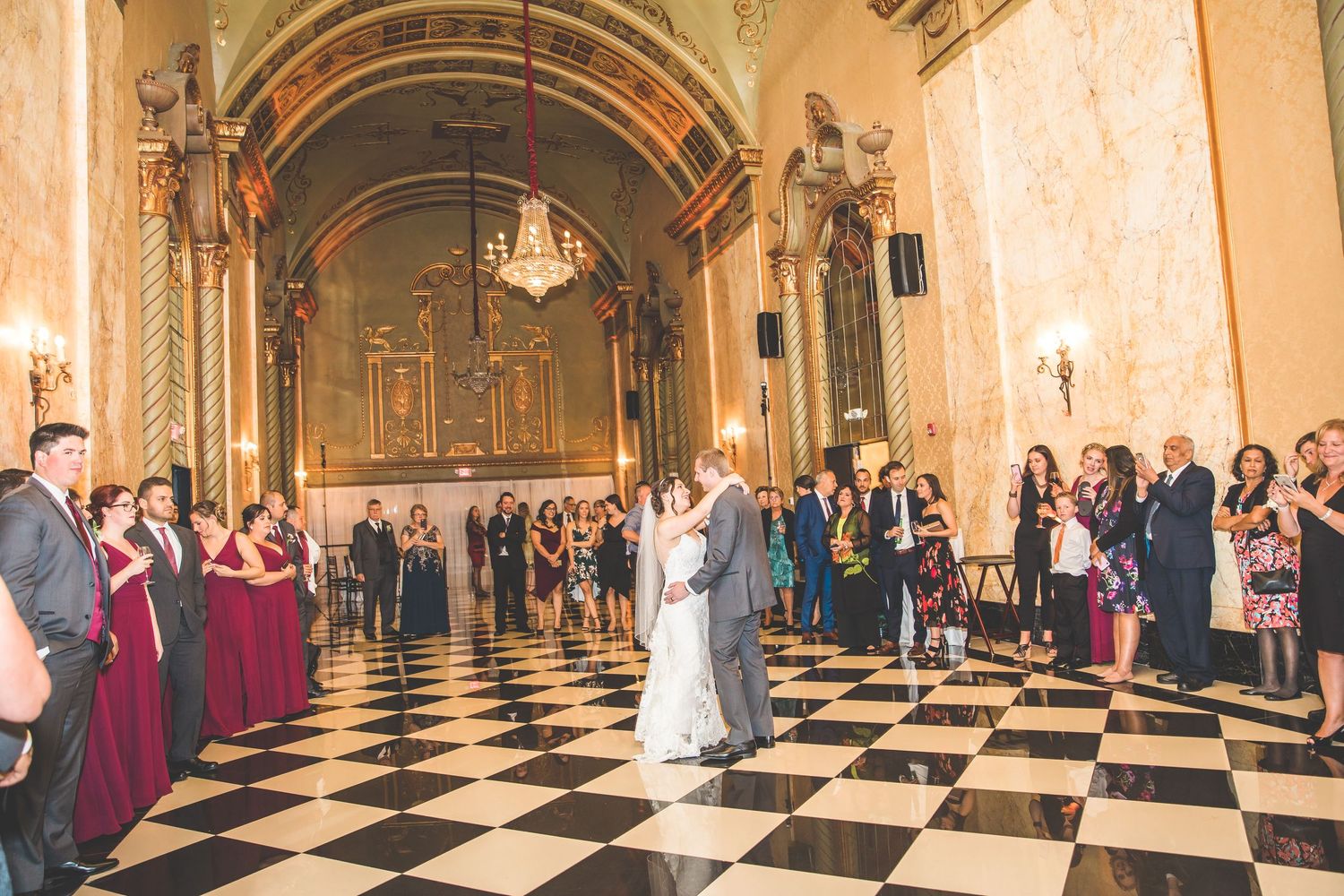 Newlyweds share their first dance in an elegant ballroom with checkered floors and ornate architecture.